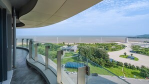 A balcony with a curved glass railing overlooking a parking area, a beach, and a coastal view.