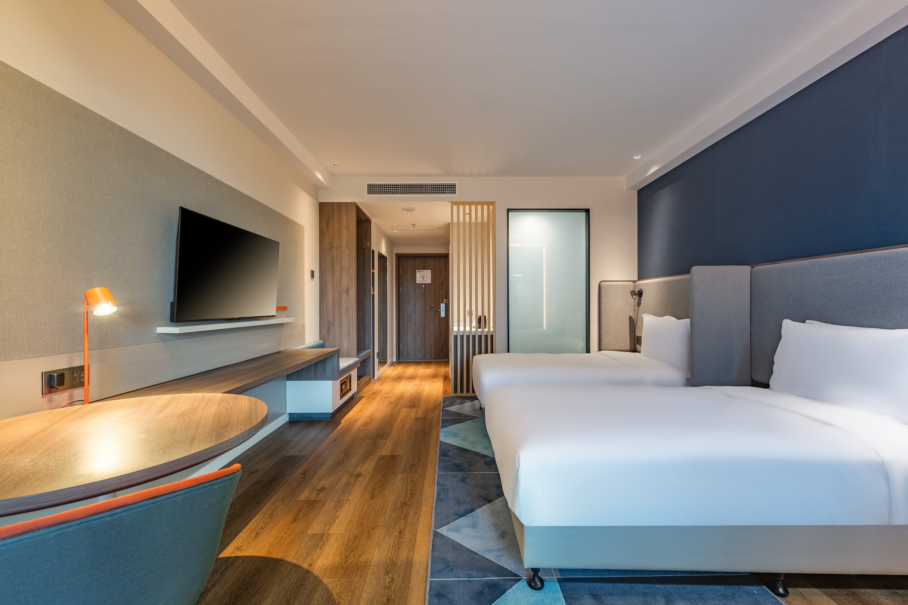 A modern hotel room with a large bed, a desk, a flat-screen TV, and a bathroom visible through a glass door.