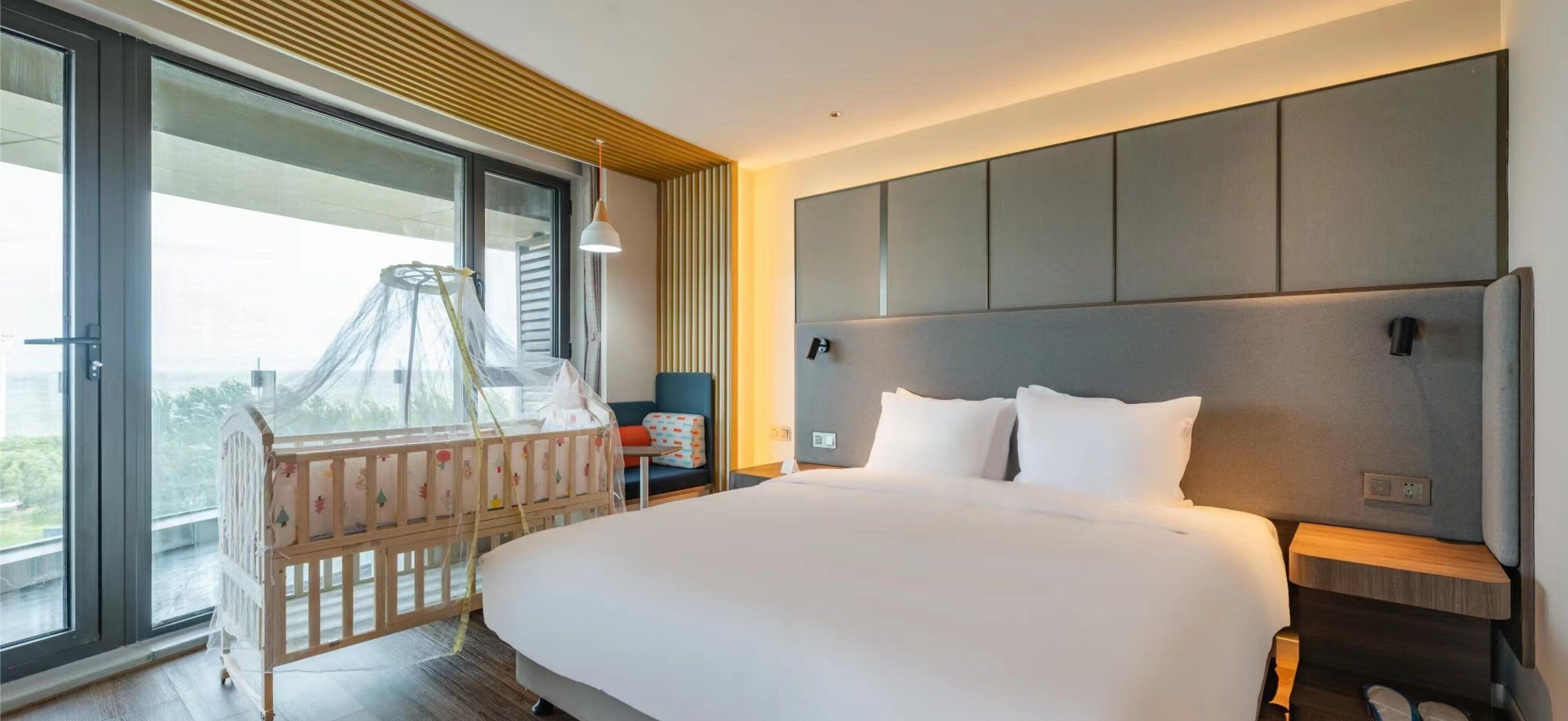 Habitación de hotel moderna con una cama grande, una mesita de noche, un buró, una ventana con vistas y una cuna.