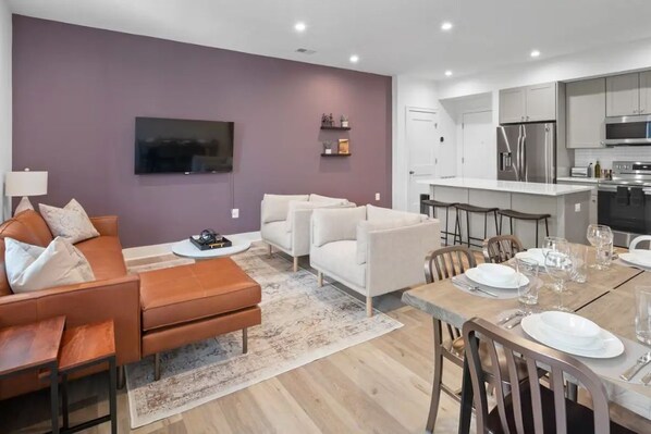 A modern living room with a sofa, coffee table, and TV. Adjacent is a dining area with a wooden table set for four. - 1/2 Block from King Street, Elegant Old Town Oasis ~ King Bed♛ ~ Free Parking (Alexandria)