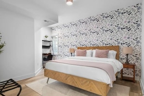 Une chambre à coucher avec un lit en bois, une table de chevet, une lampe, une chaise, un bureau et un mur recouvert d’un papier peint à motifs floraux.