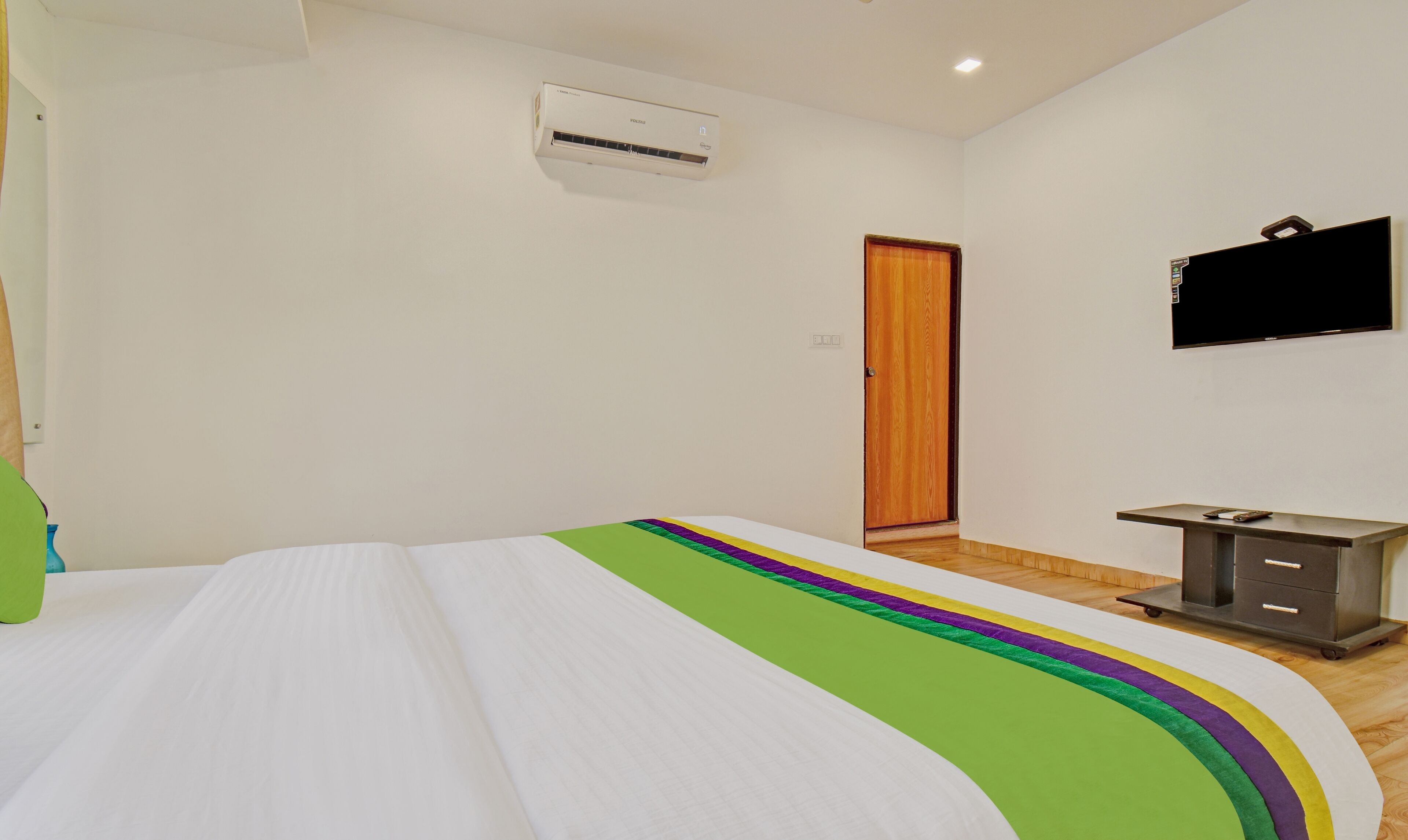 A bedroom with a bed, an air conditioning unit, a wooden door, a TV, and a small desk with drawers.