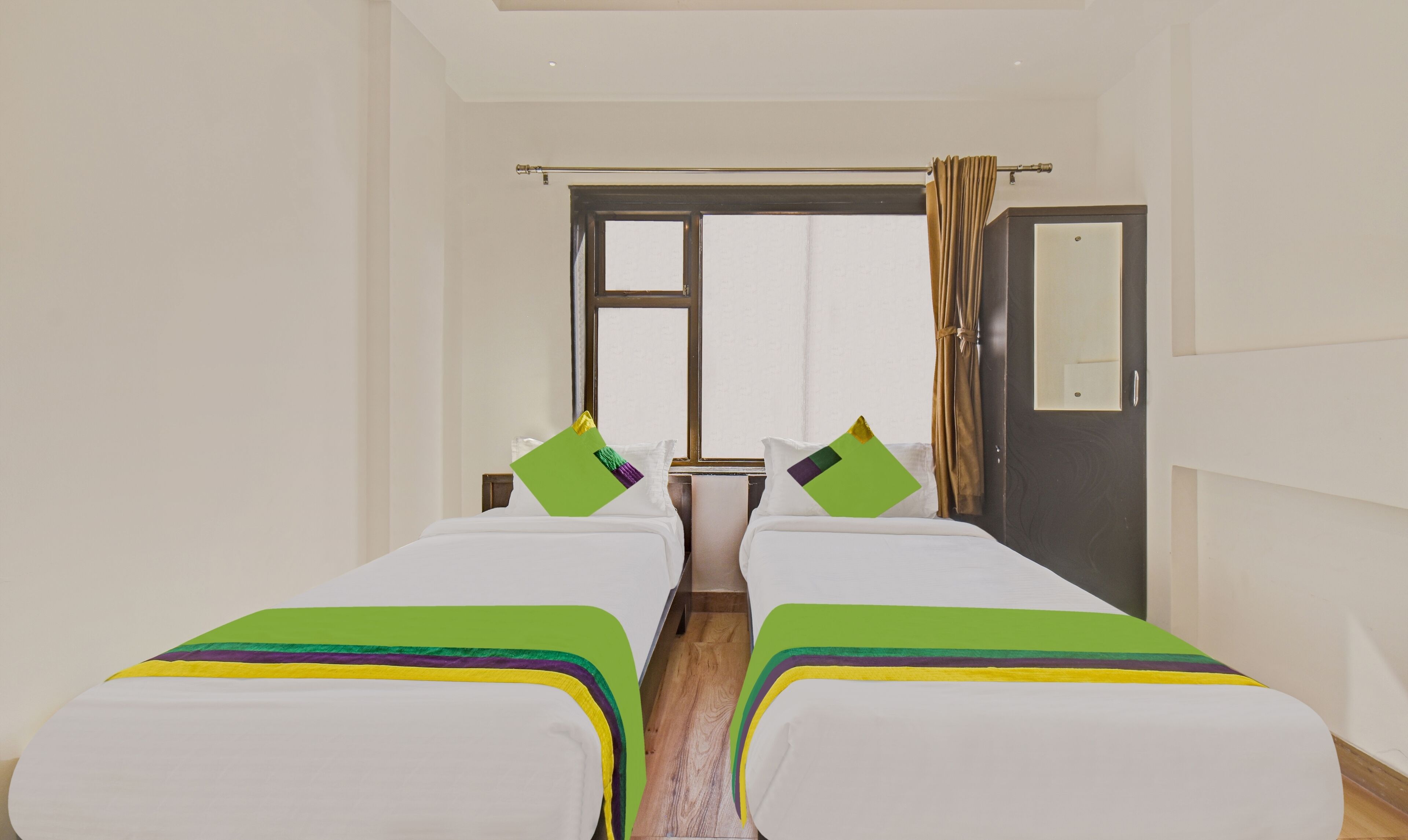 A hotel room with two single beds, a window with curtains, a wooden floor, and a door with a mirror.