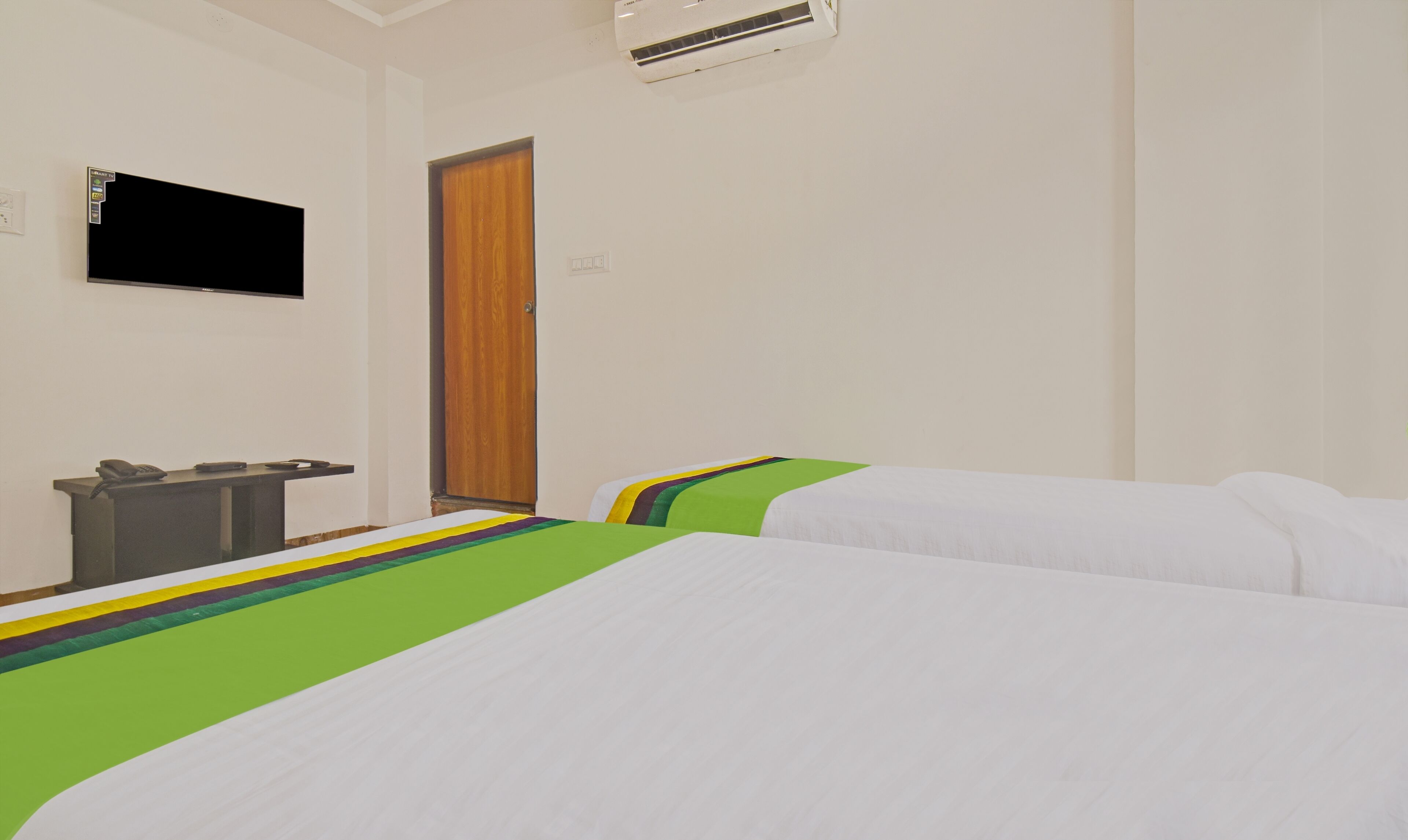 A hotel room with two beds, a desk with a telephone, a TV mounted on the wall, and an air conditioning unit.