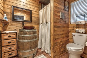 A rustic bathroom with wooden walls, a wooden barrel sink, a toilet, and a window with blinds.