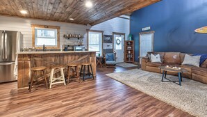 A spacious kitchen with a wooden ceiling, a bar area with stools, a living room with a sofa and a coffee table, and a blue accent wall.