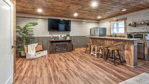 A kitchen with wooden flooring, a wooden ceiling, a large TV, and a bar area with stools.
