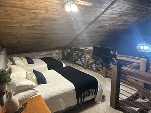 A cozy attic bedroom with a wooden ceiling, a bed with white and blue bedding, a TV, and a small table with a plant.