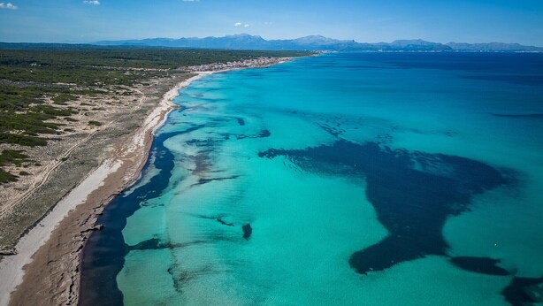 An aerial view of a coastal area with a clear beach, turquoise waters, and distant mountains.