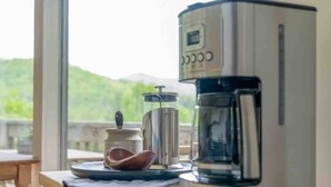 Una cafetera sobre una mesa de madera, con vistas a zonas verdes y montañas al fondo.