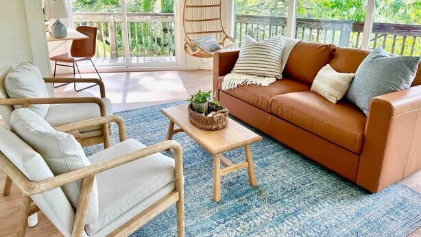 Una sala de estar acogedora con un sofá de cuero marrón, un sillón de madera y un sillón colgante. En el centro hay una mesita con una cesta y una lámpara.