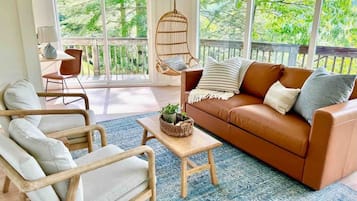 A cozy living room with a brown leather sofa, a wooden armchair, and a hanging chair. A small table with a basket and a lamp is in the center.