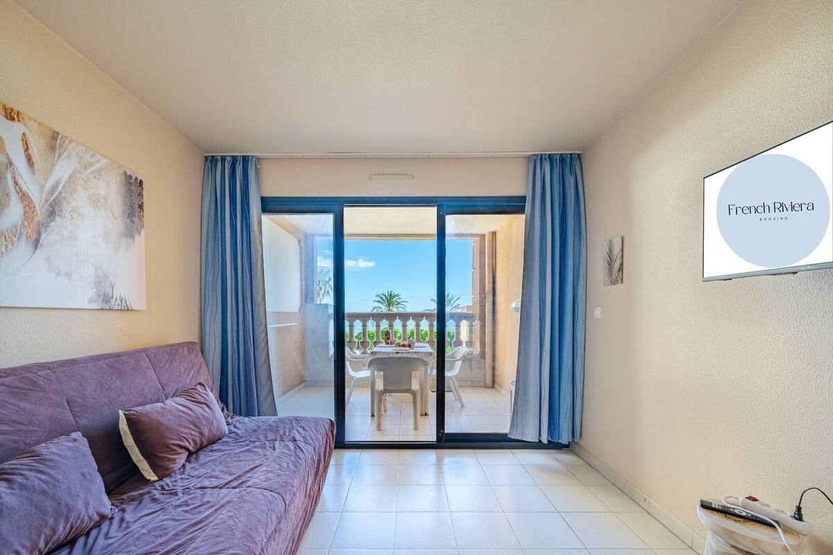 A room with a purple sofa, a sliding glass door leading to a balcony with a table and chairs, and a sign for French Riviera Booking.