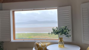 Et værelse med et stort vindue der kigger ud over en strand, et rundt bord med en vase, og en stol med en mønstret pude.