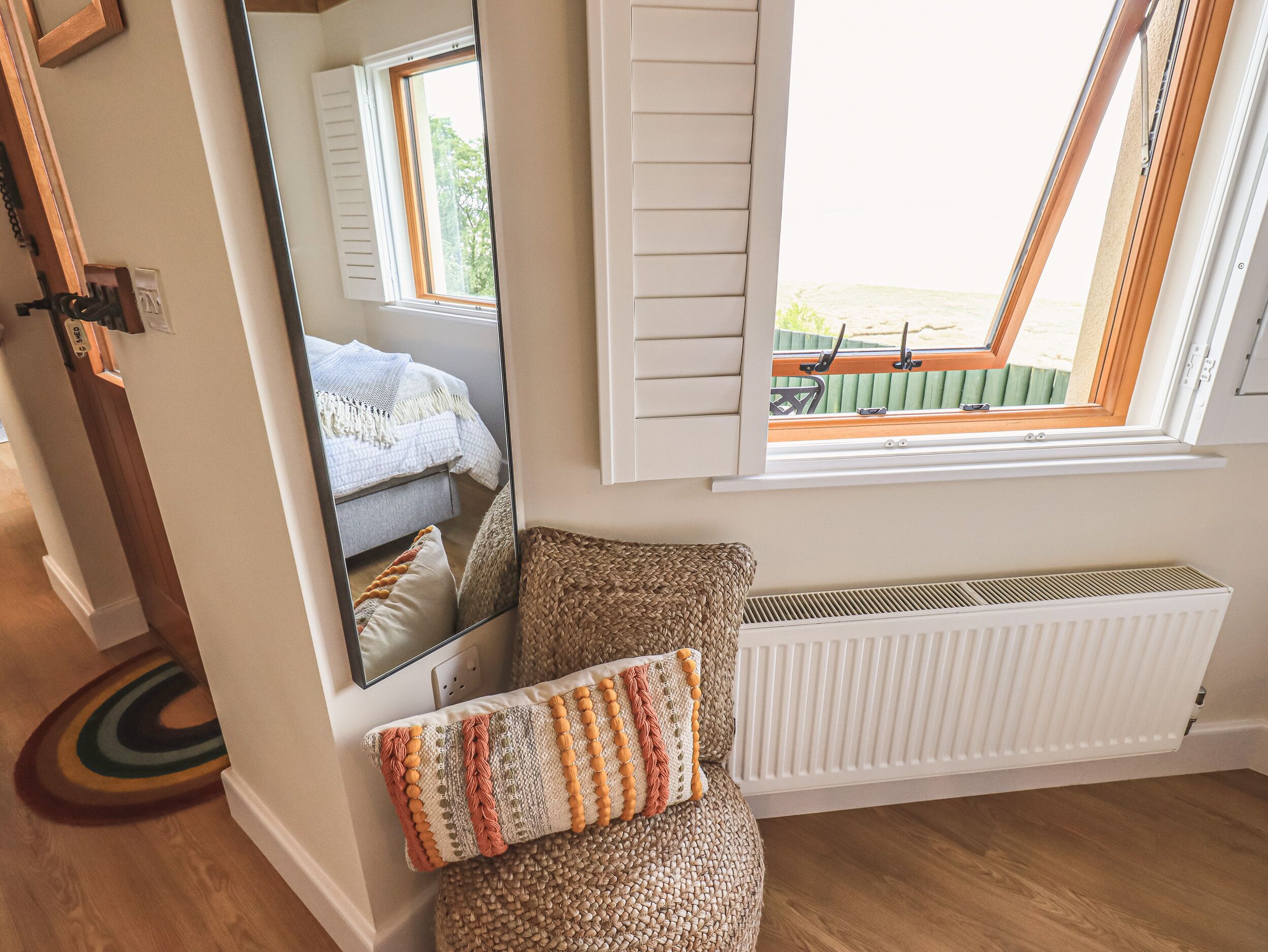 A cozy room with a large mirror, a window with shutters, a radiator, and a woven pouf with a patterned cushion.