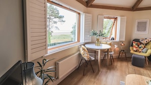 Et hyggeligt værelse med trægulv, et rundt bord og en stol. Der er vinduer med skodder, en radiator og en potteplante.