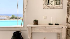 A room with a white console table, a black vase with a plant, a wall-mounted mirror, and a view of a pool and landscape. - Villa Ramona in Mykonos (Mykonos)
