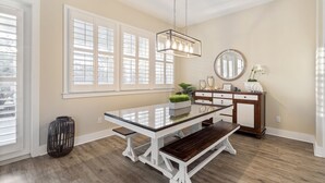 Uma área de refeições com mesa de vidro, bancos de madeira, luminária pendente, espelho e uma planta em um armário de madeira.