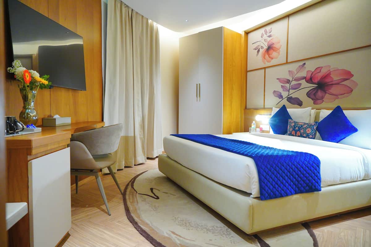 A hotel room with a bed, a desk, a chair, a TV, and a large floral painting on the wall.