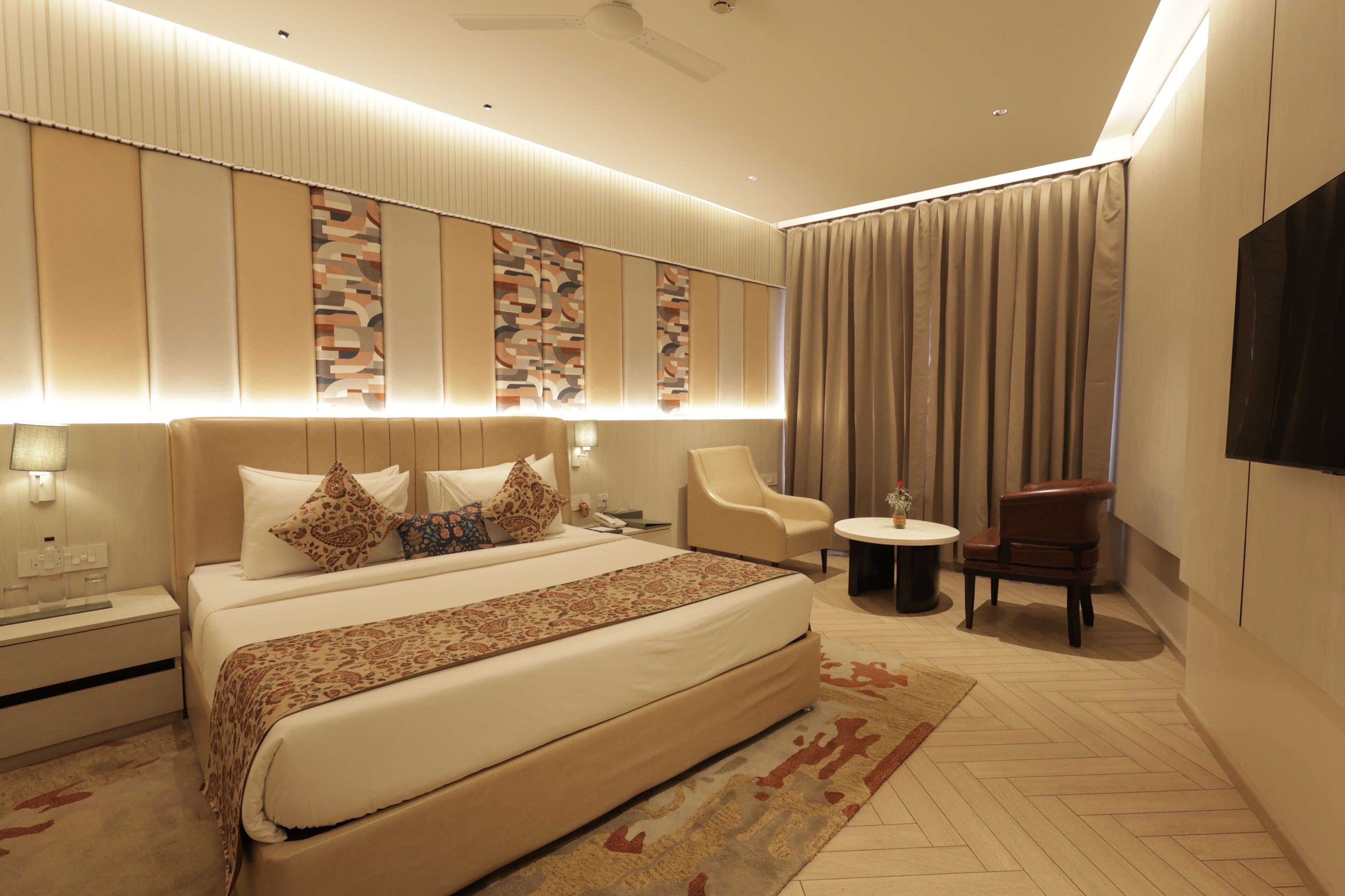 A hotel room with a large bed, a small round table, a chair, a TV, and a ceiling fan.