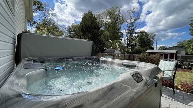 Un bain à remous doté d’un panneau de commande, entouré d’un jardin avec des arbres et une maison en arrière-plan.