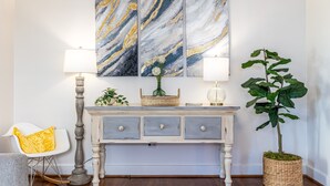 Une pièce avec une console blanche, deux lampes fixées au mur, un grand tableau abstrait et une plante en pot.