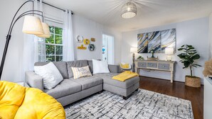 Un salon moderne avec un canapé d’angle gris, un fauteuil poire jaune, un tapis à motifs et une grande fenêtre ornée de rideaux légers.
