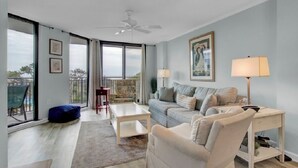 A spacious living room with a sofa, coffee table, and a desk with a lamp. There are large windows and a balcony visible. - Award-Winning Resort Amenities | Shipwatch 210B (Isle of Palms)