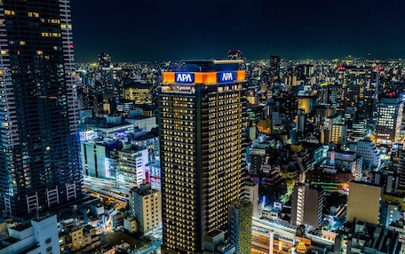 A high-rise building with APA signage, surrounded by a dense urban area with various other buildings and illuminated streets.