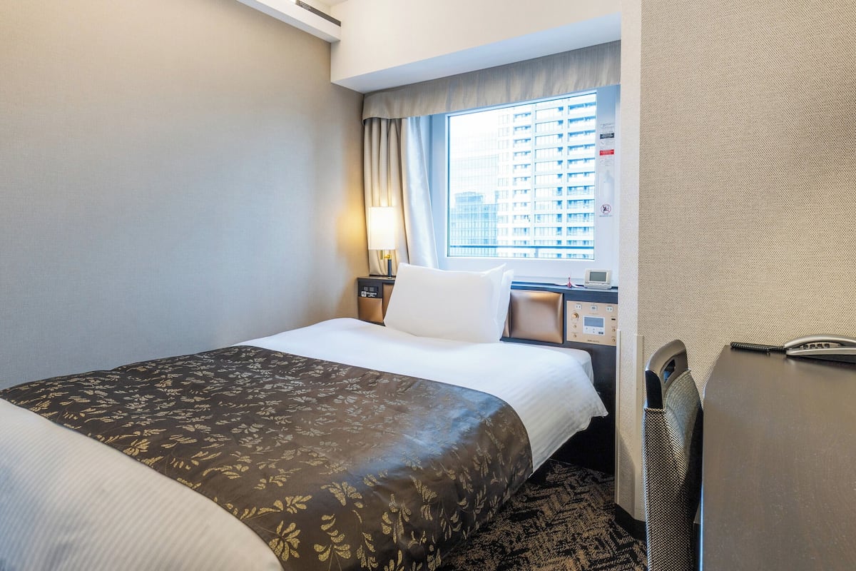 A hotel room with a bed, a desk, a lamp, a telephone, and a window with a city view.