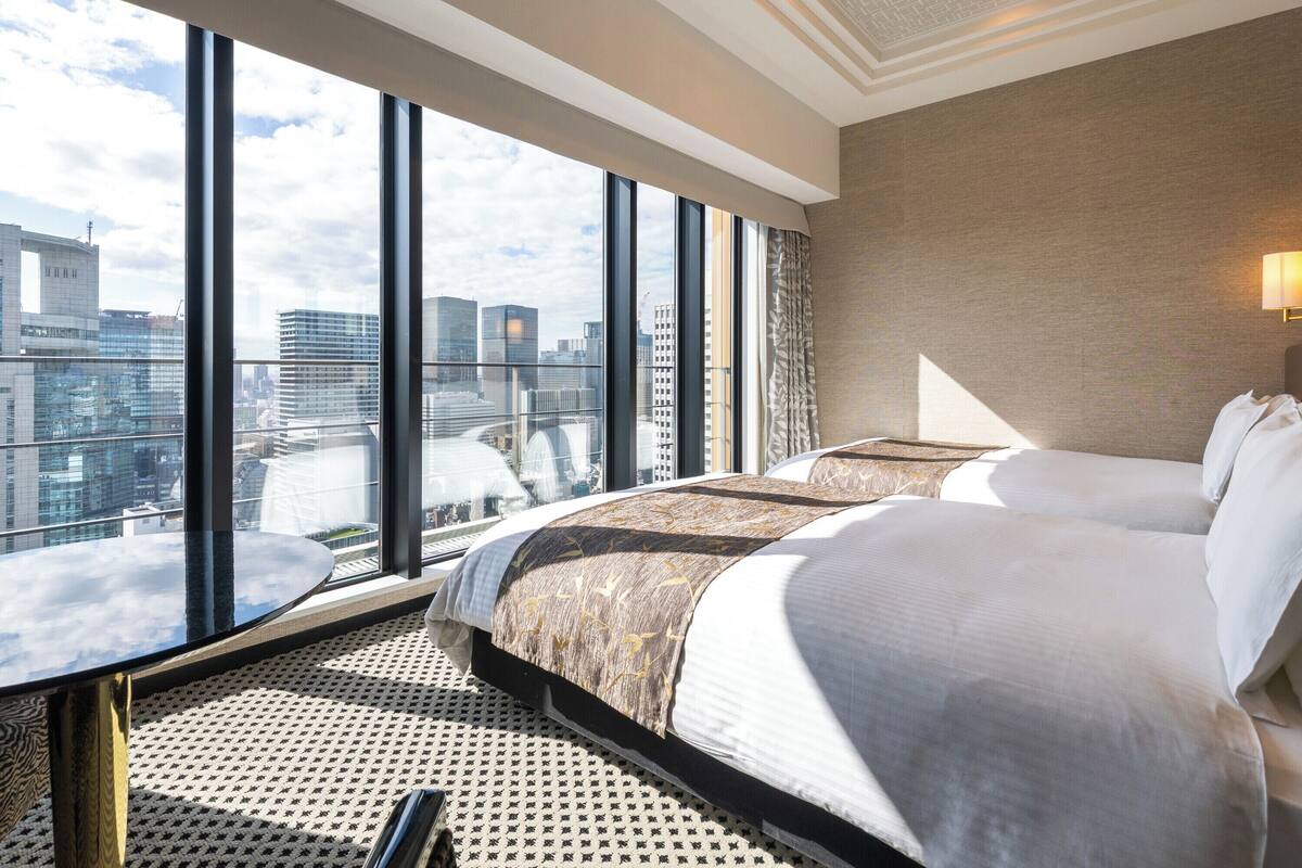 A hotel room with a large window offering a city view, a bed with patterned bedding, and a round table with a remote control.