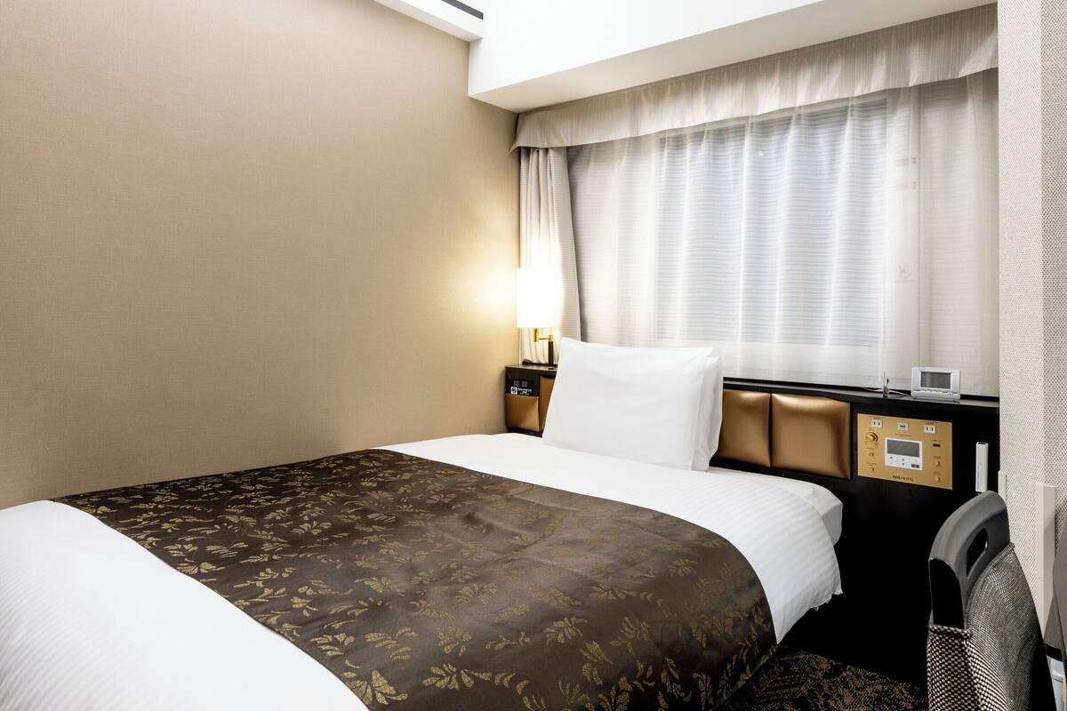 A hotel room with a large bed, a nightstand with a lamp, a control panel, and a window with curtains.