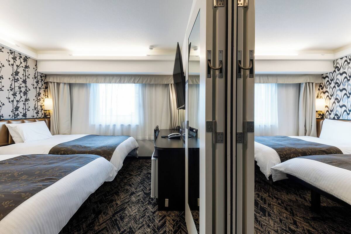 A hotel room with two beds, a desk, a TV, and a shower area.