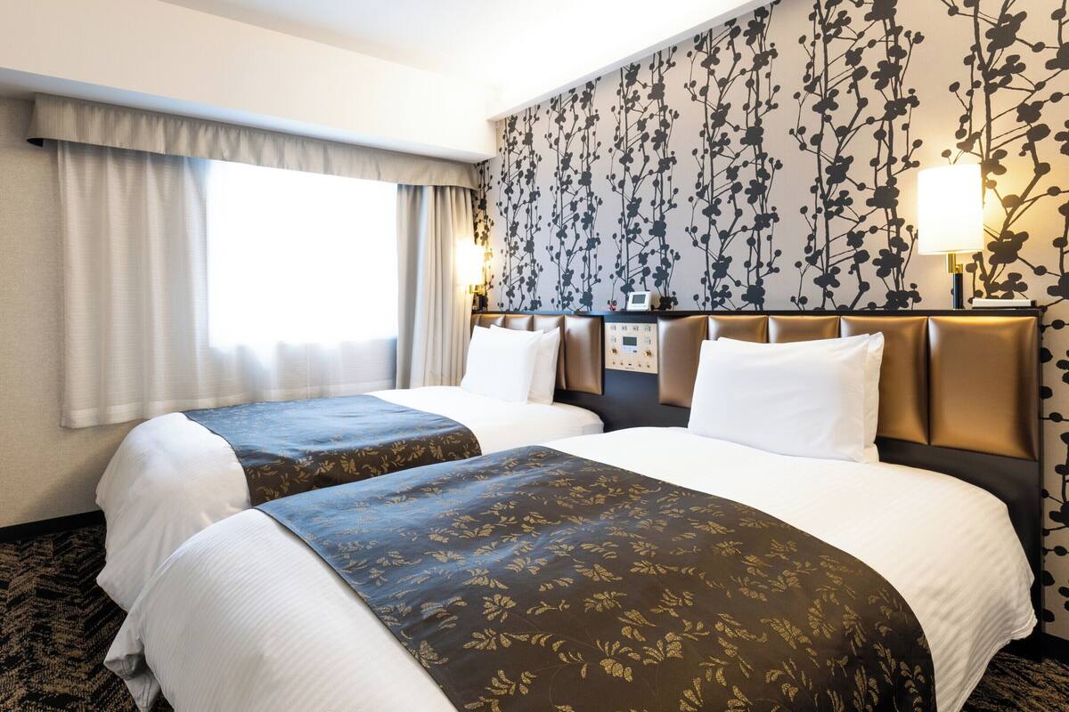A hotel room with two beds, a large window with curtains, a floral wallpaper, and a bedside table with a lamp.