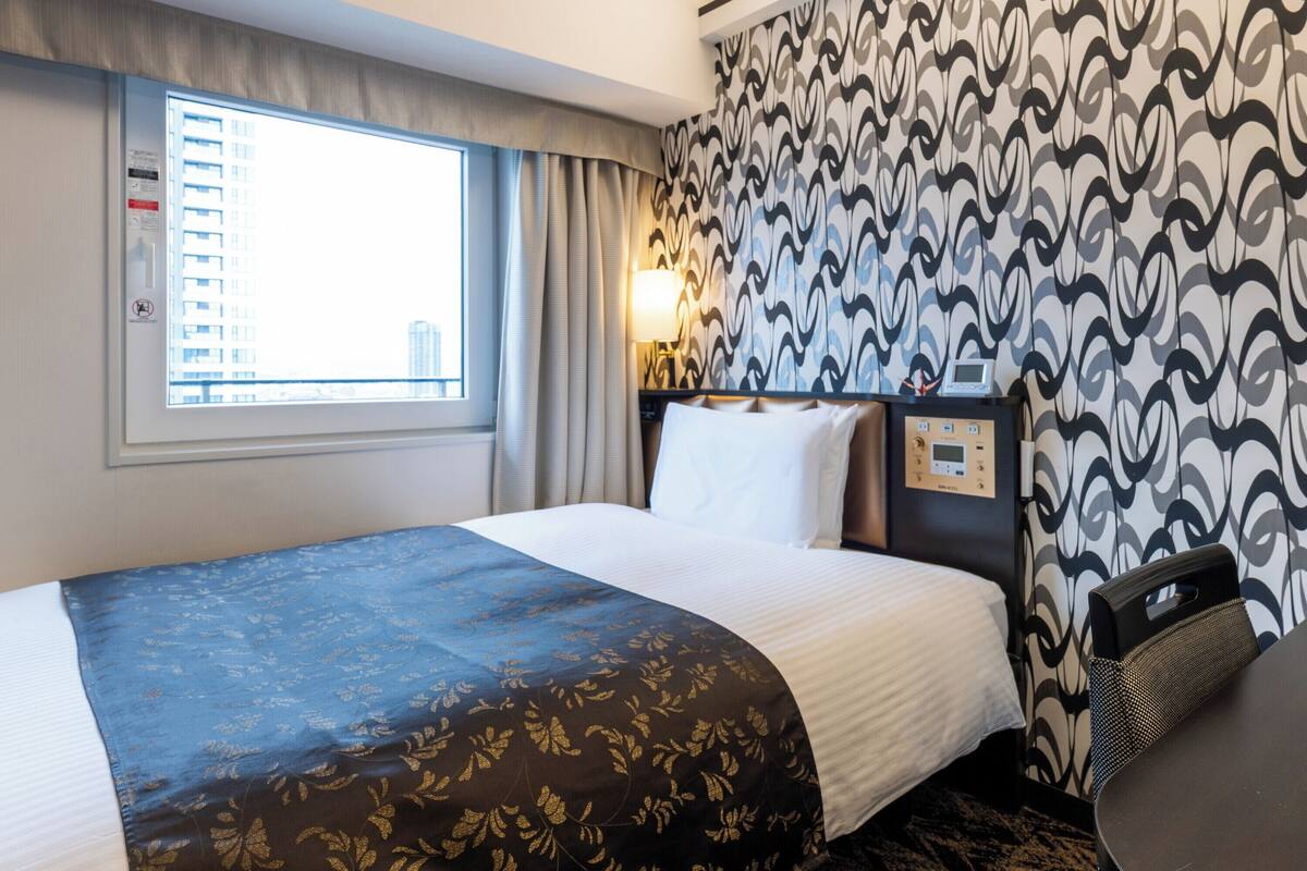 A hotel room with a large bed, a window with curtains, a patterned wall, and a control panel on the wall.