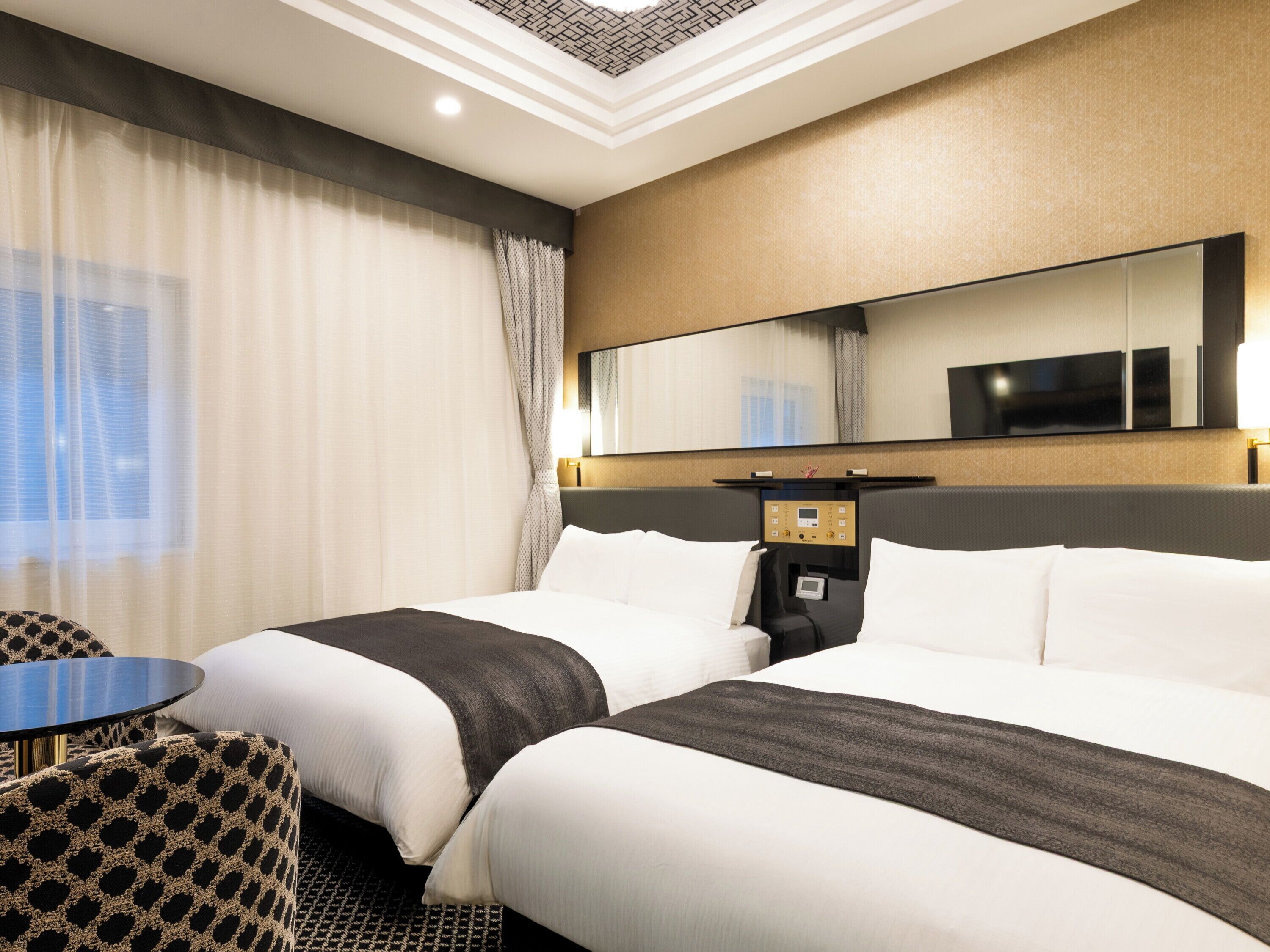 A hotel room with two beds, a desk, a chair, a large mirror, and a control panel.