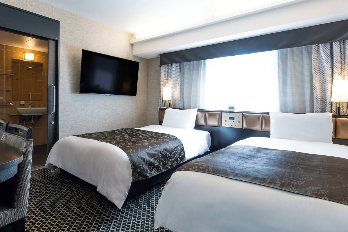 A hotel room with two beds, a TV mounted on the wall, a desk with a chair, and a window with curtains.