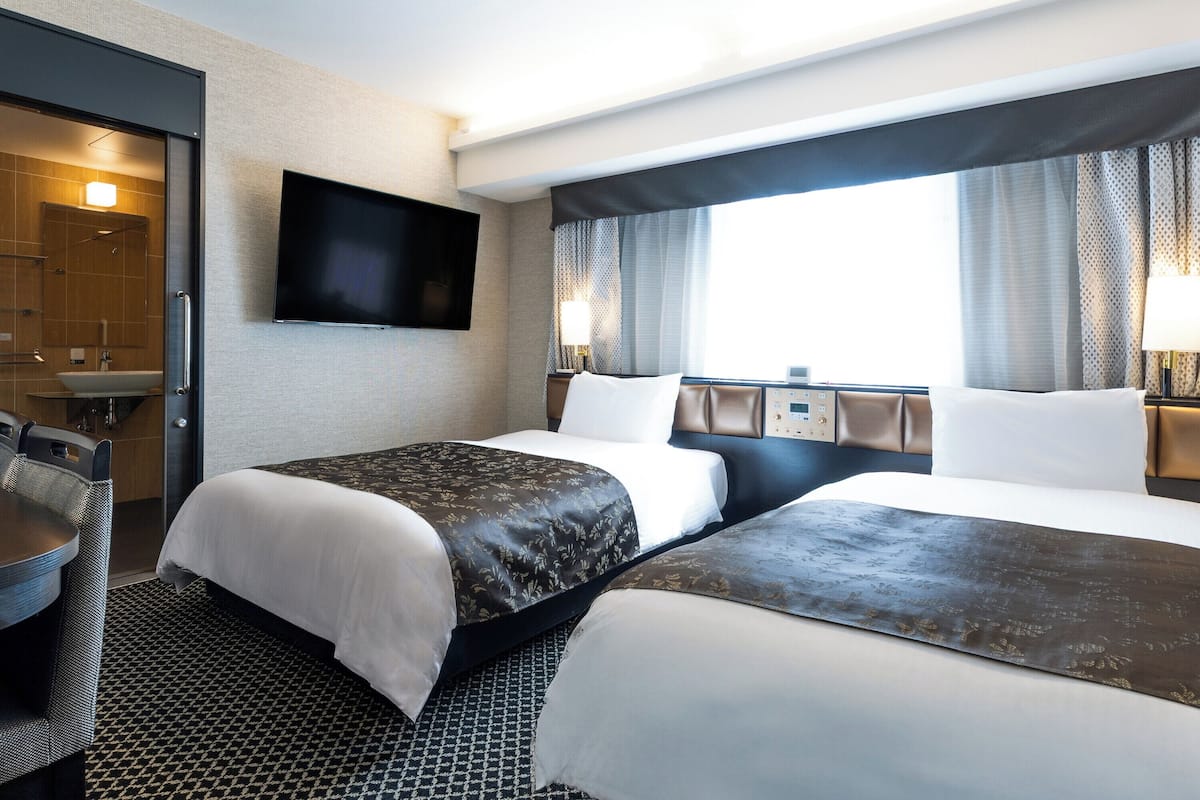 A hotel room with two beds, a TV mounted on the wall, a desk with a chair, and a window with curtains.