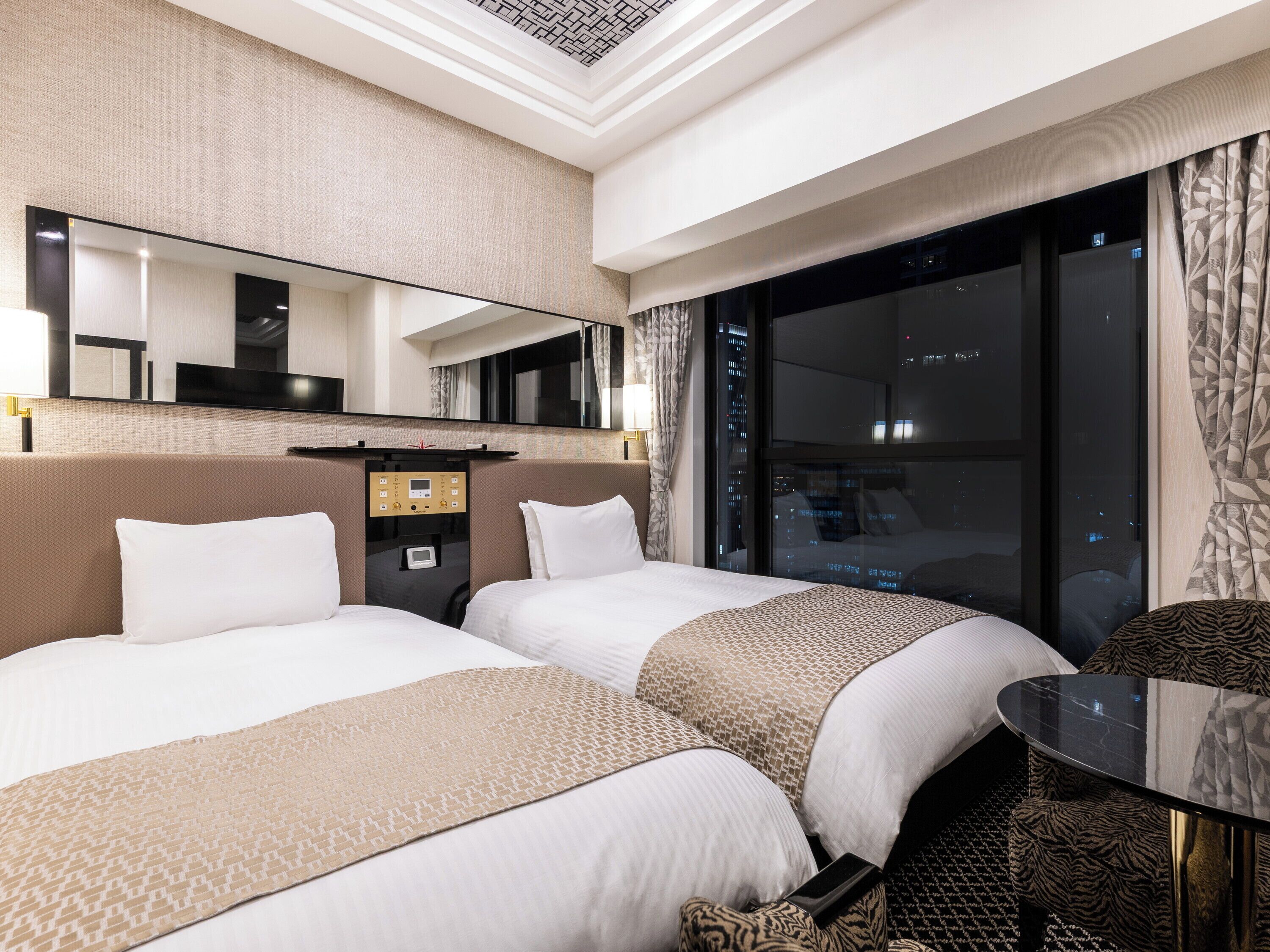 A hotel room with two beds, a large mirror, a control panel, a window with a city view, and a small table.