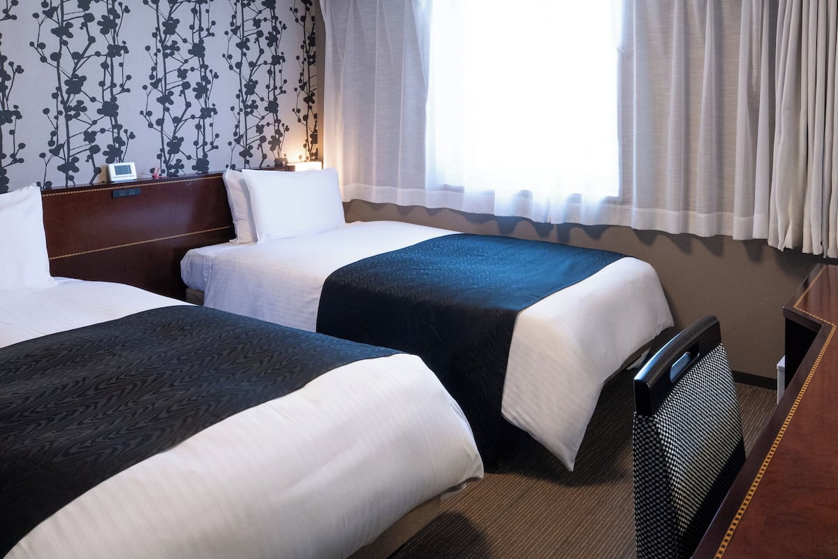 A hotel room with two beds, a wooden headboard, a bedside table with a lamp, a window with sheer curtains, and a floral wallpaper pattern.