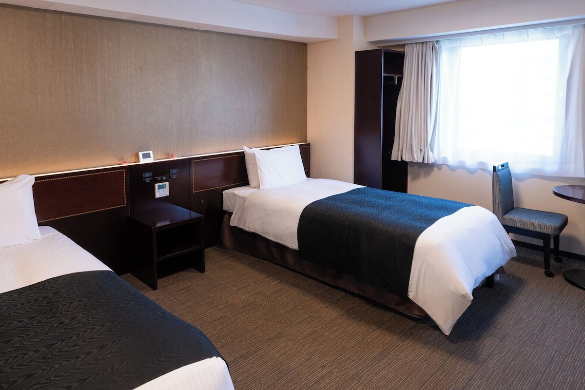 A hotel room with two beds, a desk, a chair, and a window with curtains.