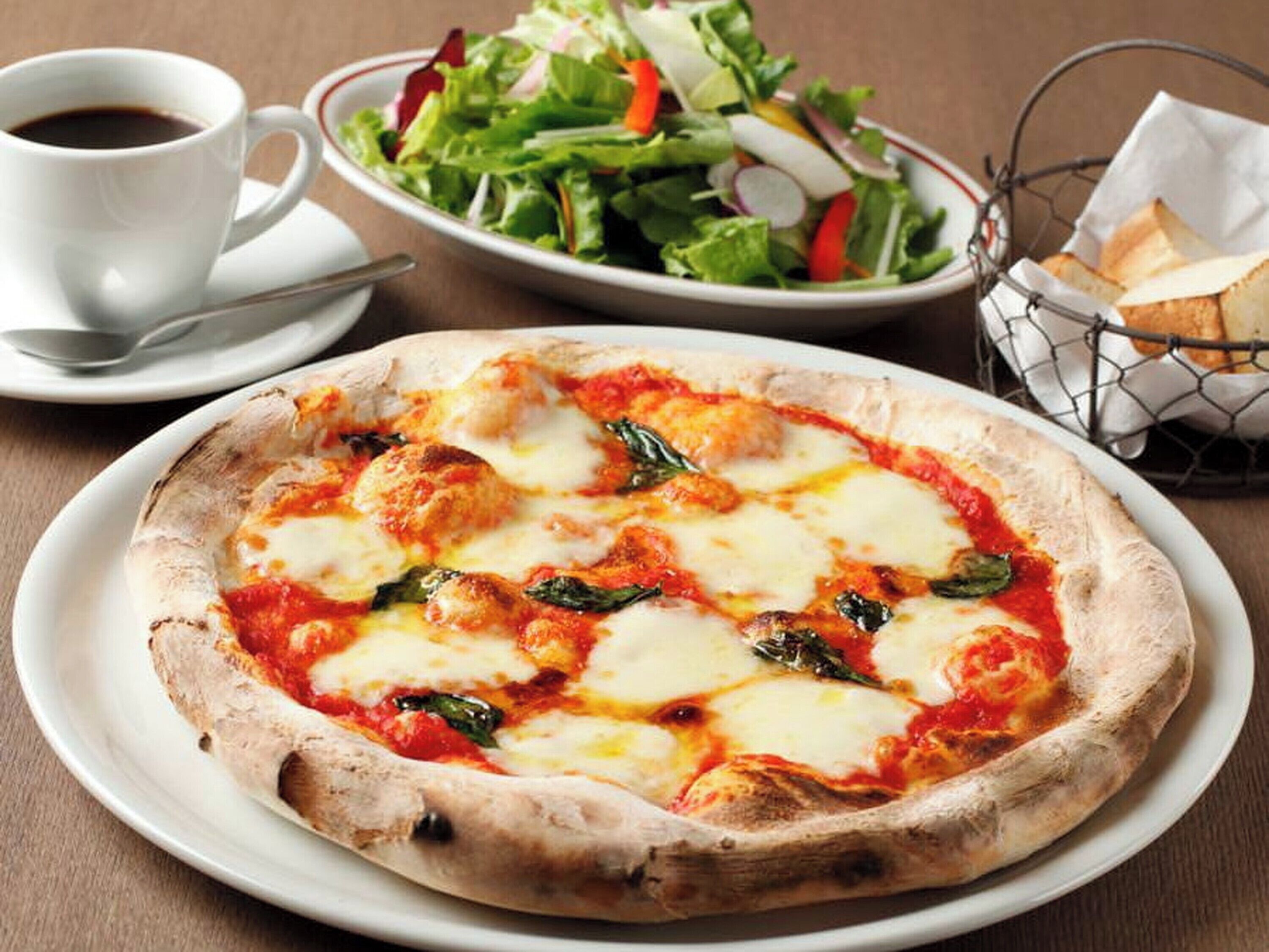 A pizza with cheese and spinach on a white plate, a cup of coffee, a salad, and a basket of bread on a table.