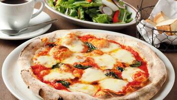 Une pizza à la mozzarella et aux épinards, servie sur une assiette blanche, une tasse de café, une salade et un panier de pain, le tout posé sur une table.