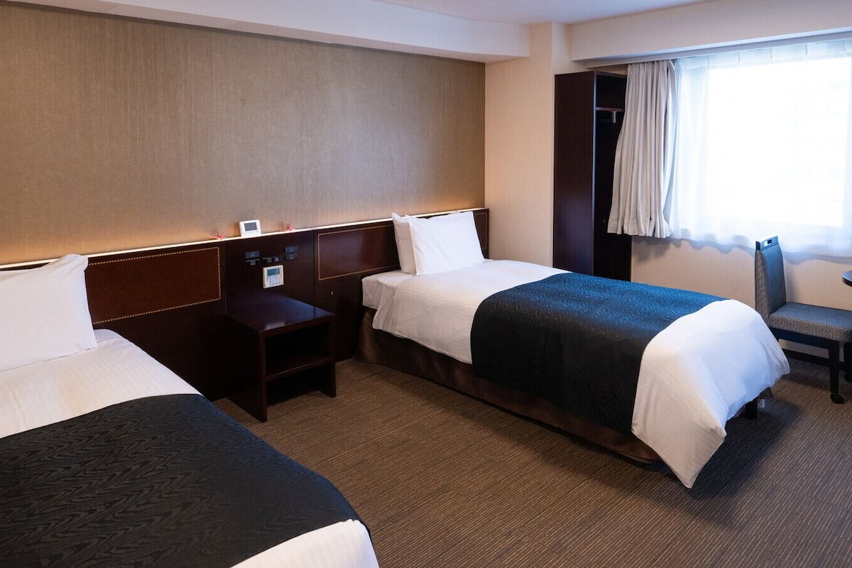 A hotel room with two beds, a desk, a chair, and a window with curtains.