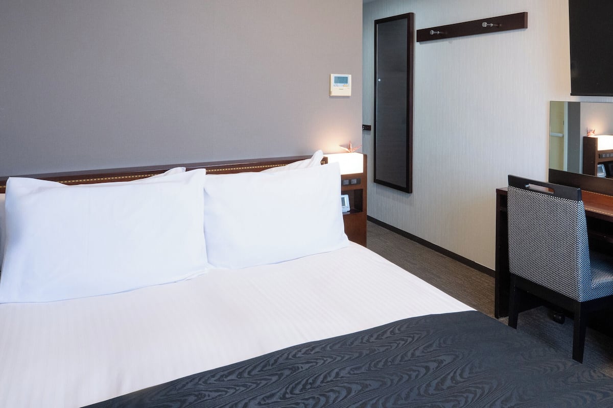 A hotel room with a bed, a desk, a chair, a nightstand, a lamp, and a wall-mounted TV.