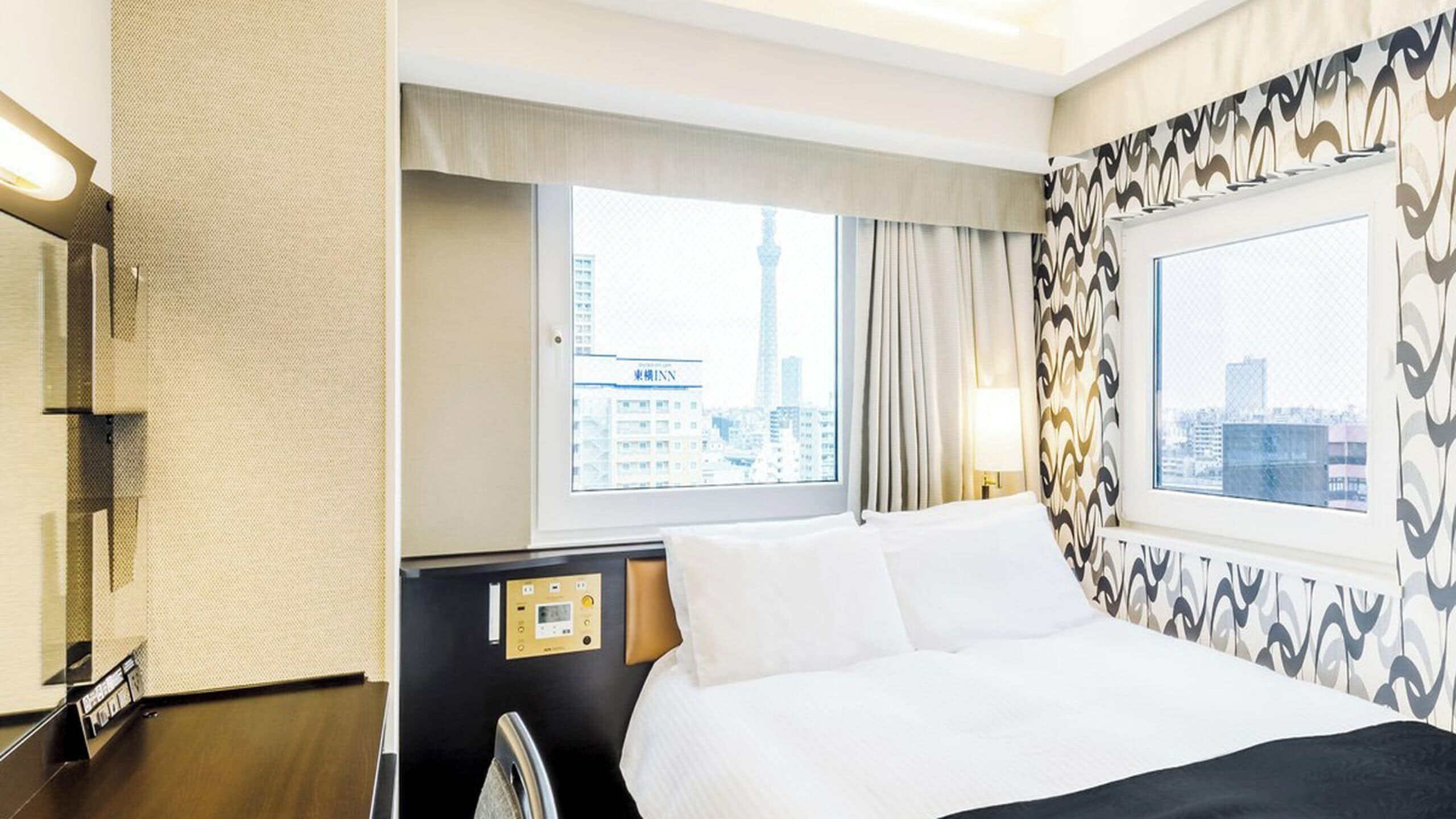 A hotel room with a bed, a control panel, a window with a city view, and patterned curtains.