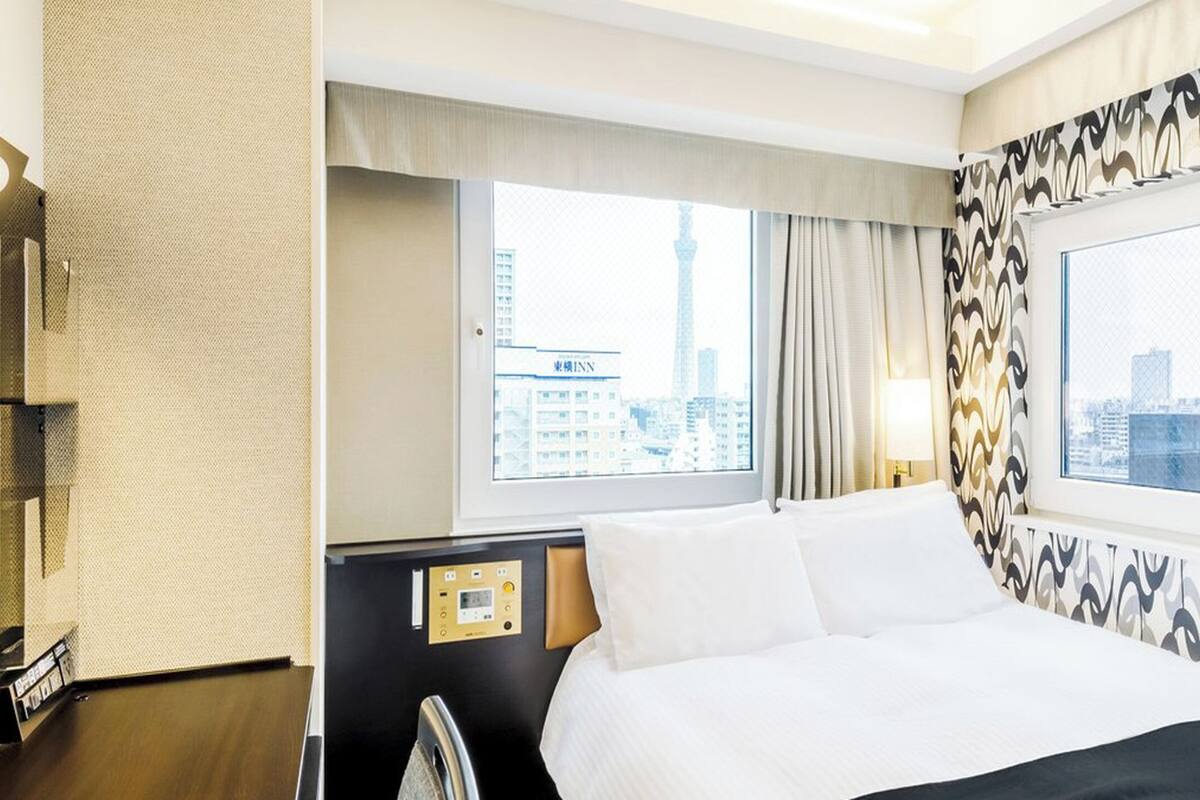 A hotel room with a bed, a control panel, a window with a city view, and patterned curtains.