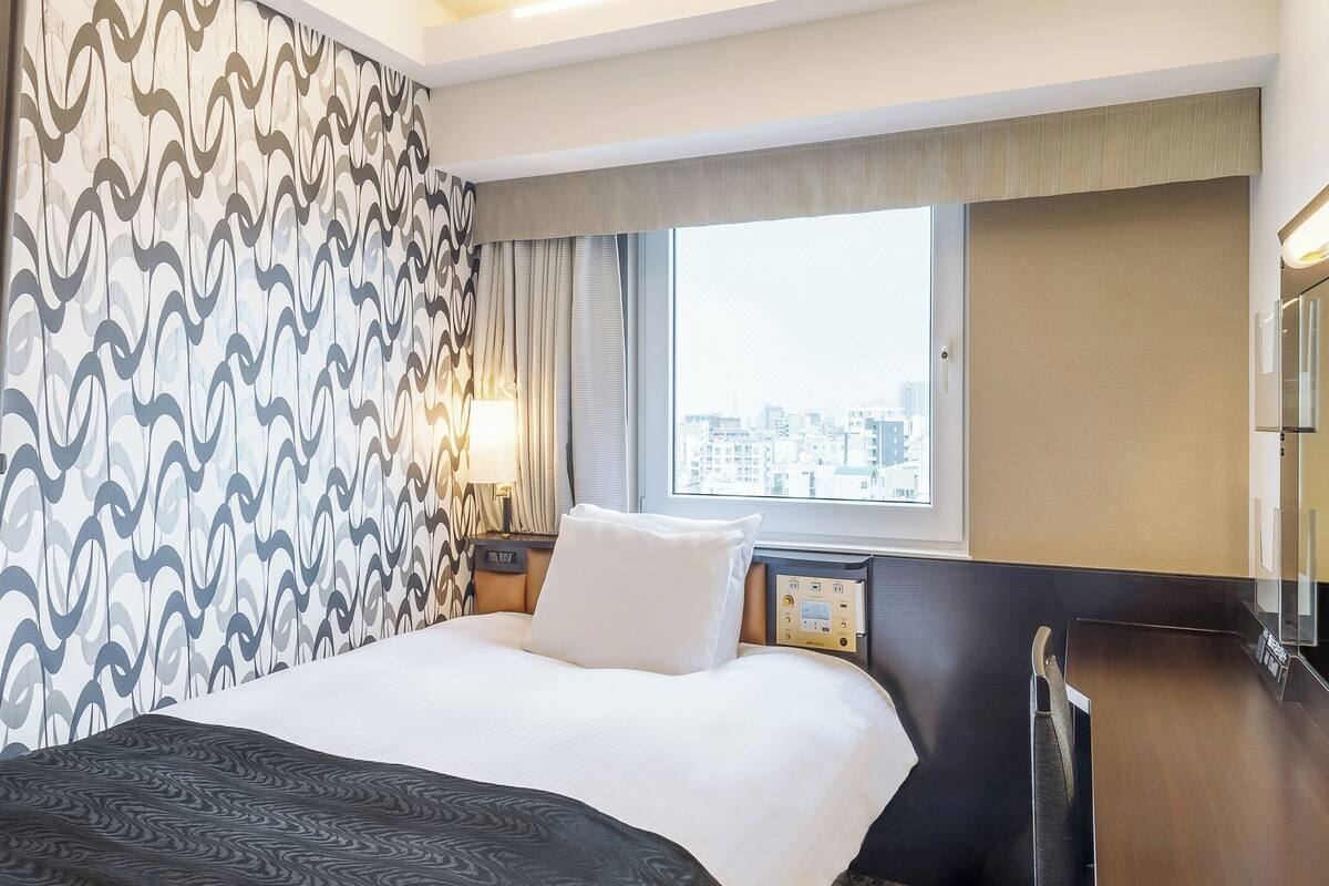 A hotel room with a bed, a nightstand, a lamp, a window with a city view, and a minibar.