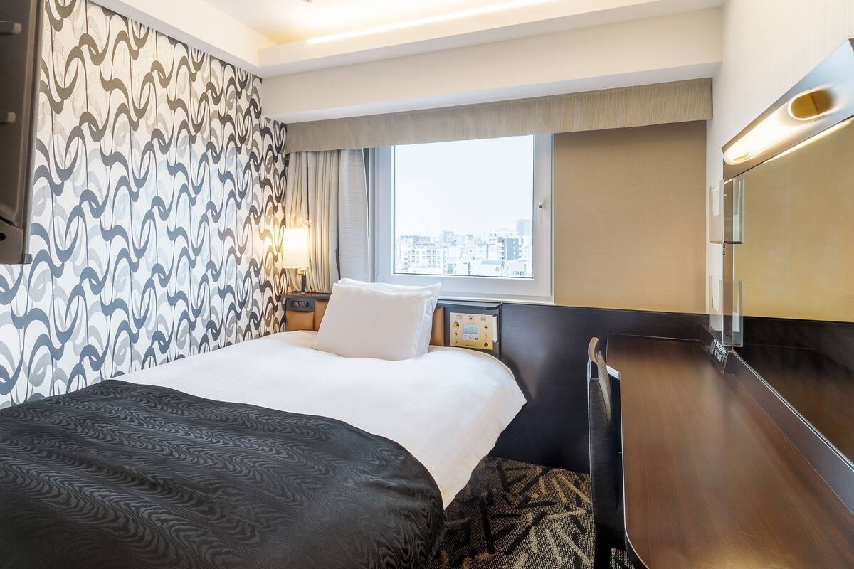A hotel room with a bed, a desk, a window with a city view, and a patterned wall.
