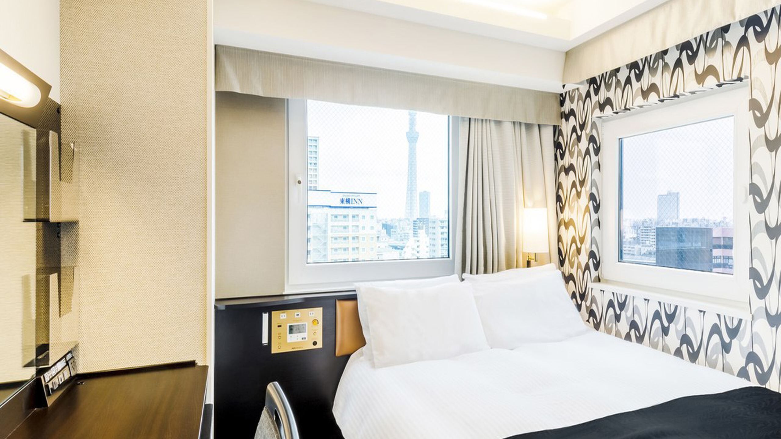 A hotel room with a bed, a control panel, a window with a city view, and patterned curtains.